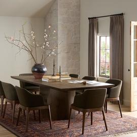 Modern rustic dining table set with dark wood chairs, large vase centerpiece, and plush rug in warmly lit room.